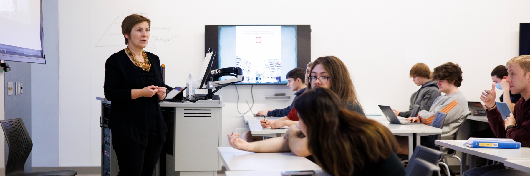 Instructor standing at the front of the classroom and speaking to rows of students at desks.