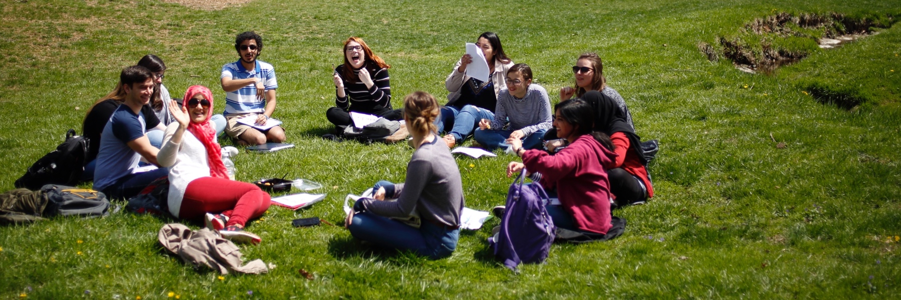 A group of IU students sit in a circle on a grassy lawn, some laughing and smiling at the camera