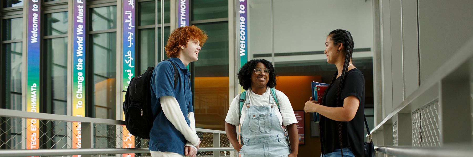 Three students, one male and two female, talking in the School of Global and International Studies building with brightly colored signs in the background welcoming students in various languages.