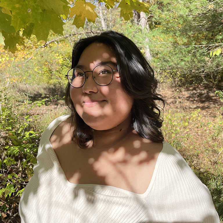 Wren Garcia, a woman with glasses and long dark hair, standing in a grove of trees and looking up, toward the sky.