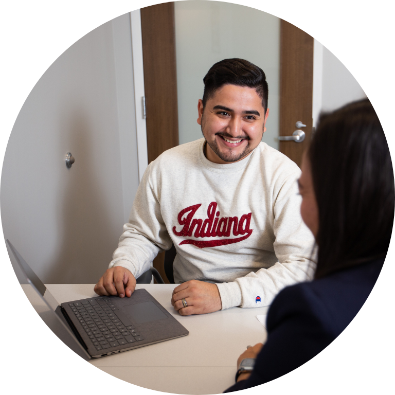 Smiling individual in an Indiana University sweatshirt sitting with a laptop and looking across a desk.