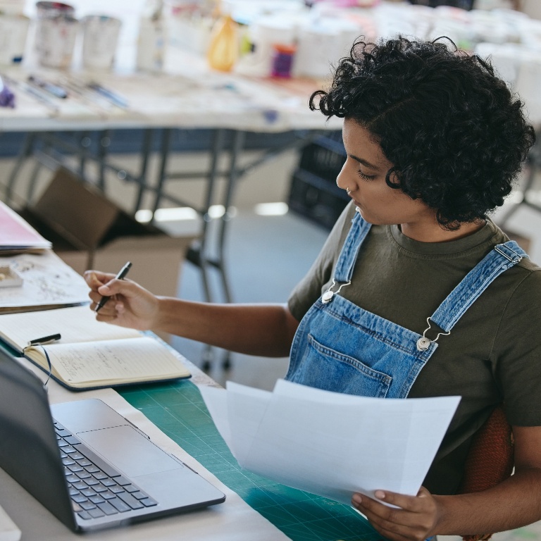 Woman standing at a desk, working with a laptop and writing on a notepad.