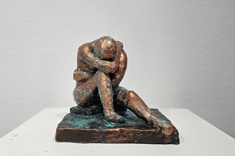 Small ceramic sculpture by Aspen Lyons titled Therapy II. A rustic human figure with legs outstretched and arms wrapped around its body. The piece is mostly cold colored with some green oxidation on its base.