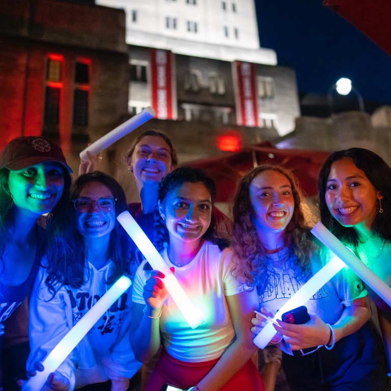 a group of students hold glow sticks in front of a lit up IMU at night