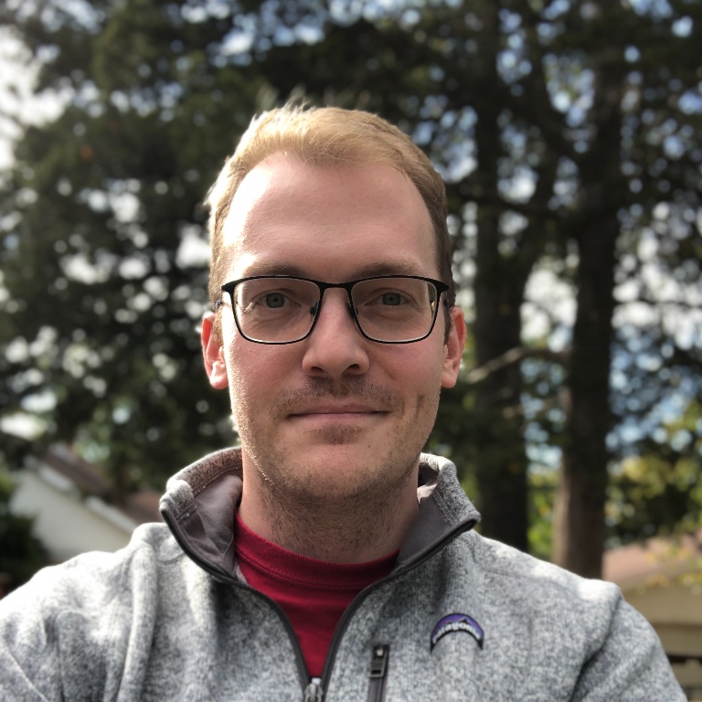 Clinton Roeth-McKay staff photo. White man in his thirties, wearing a grey pullover, smiling with blurry trees in the background.