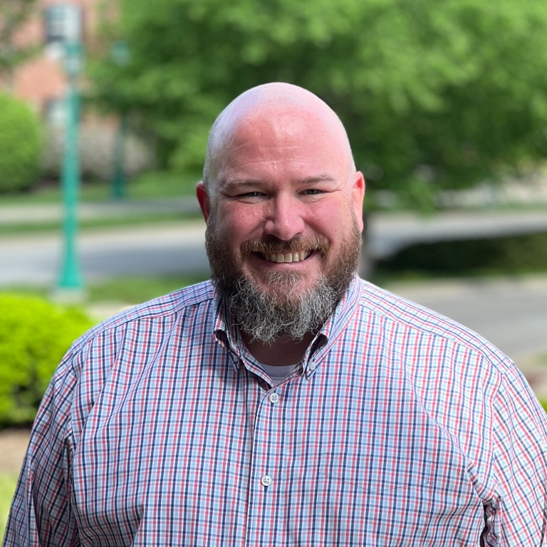 James Audley staff photo. White man with no hair wearing a plaid shirt, the IU Bloomington campus is in the background.