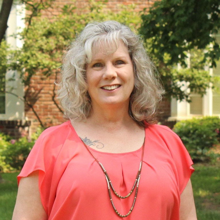 Jennifer Wentz-Montgomery staff photo. A white woman with grey hair and wearing a salmon colored top and a necklace. She is smiling with brick building and trees in the background.