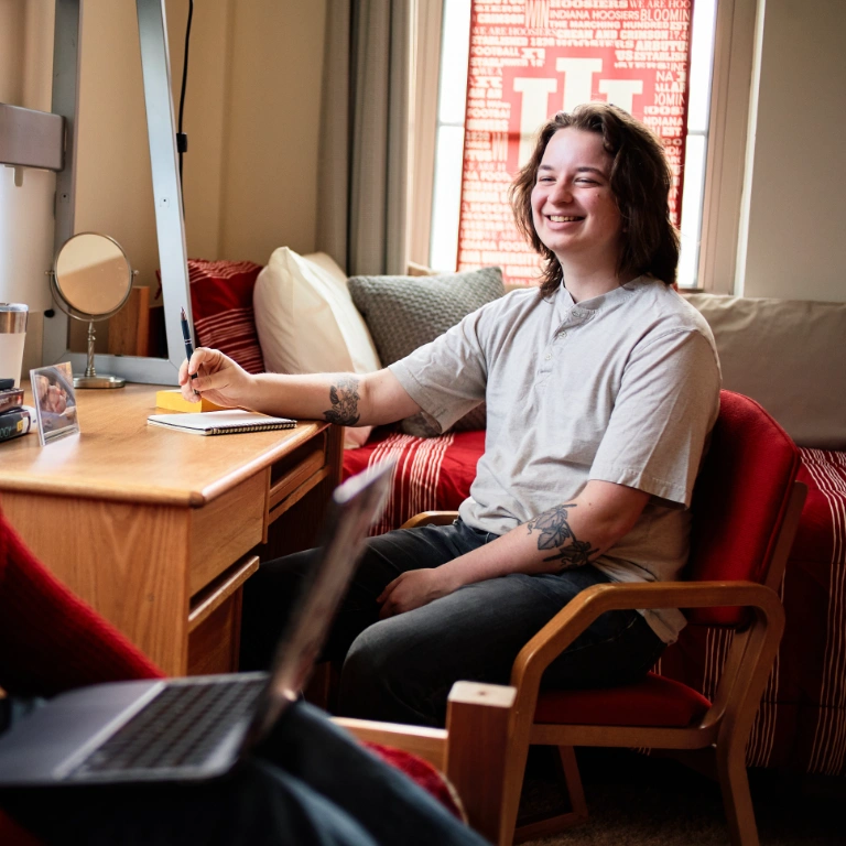 A student sits at a desk and smiles in an IU-decorated dorm room