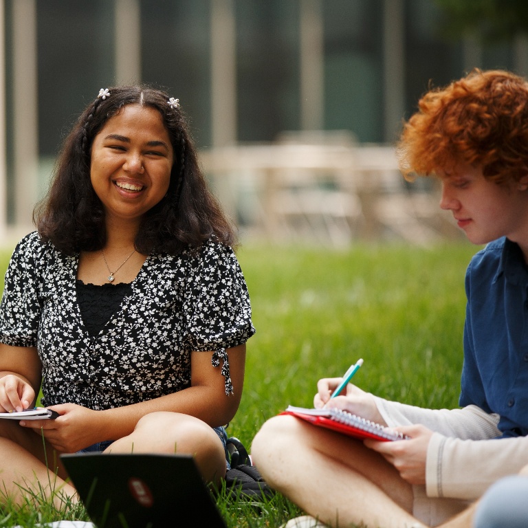 Two students working while sitting in grass outside.