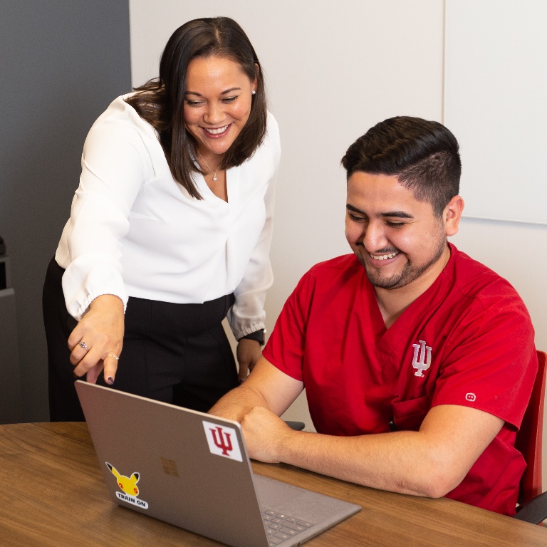 Student wearing IU scrubs and working on a laptop with an instructor assisting.